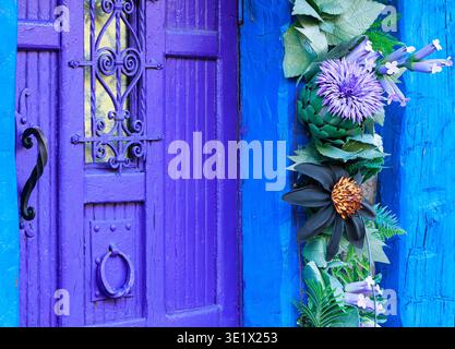 Una suggestiva porta in legno viola presenta intricati dettagli in ferro battuto ed e' adornata da una cascata di fiori artificiali e vegetazione Foto Stock