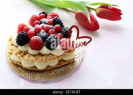 San Valentino. Deliziosa crostata di frutta a forma di cuore decorata con frutti di bosco freschi e zucchero a velo. Decorazioni a forma di cuore e tulipani rossi. Foto Stock