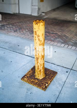 Ripresa ad alto angolo di un paletto in metallo giallo arrugginito in piedi su un marciapiede urbano in cemento con sfondo in mattoni. Foto Stock