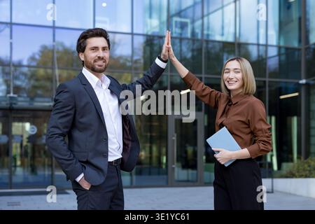 Due professionisti del business multietnici sorridenti che celebrano la collaborazione e la partnership con un gesto di cinque persone all'esterno di un moderno edificio di vetro, simboleggiando il lavoro di squadra e il successo Foto Stock