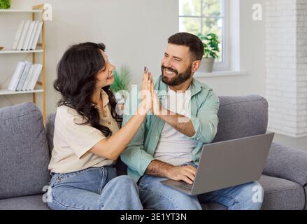 Coppia che festeggia il pagamento con il laptop a casa, cinque in alto sul divano Foto Stock