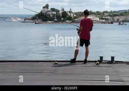 Un uomo sta su un molo vicino all'acqua, a pescare. Indossa una camicia nera e pantaloncini neri. Foto Stock