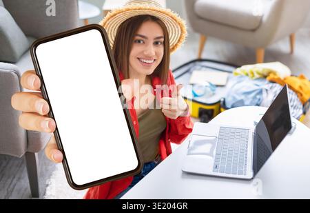 Una giovane donna mostra lo smartphone mentre è seduta alla scrivania, preparandosi per il lavoro online in ufficio domestico Foto Stock