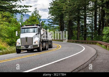 Semi-autocarro di grandi dimensioni con punta Day Cab bianca per impieghi industriali con due rimorchi ribaltabili carichi che percorrono le tortuose strade di montagna con alberi sul Foto Stock