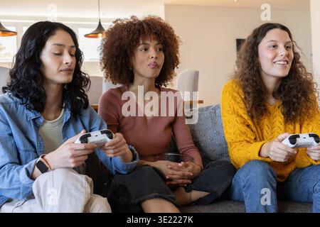 Diverse amiche sedute su un divano grigio che regge controller di gioco bianchi e guarda l'azione di gioco Foto Stock