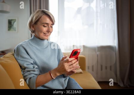 Bella donna di mezza età seduta comodamente sul divano e trascorrendo del tempo a scorrere sullo smartphone online. Foto Stock
