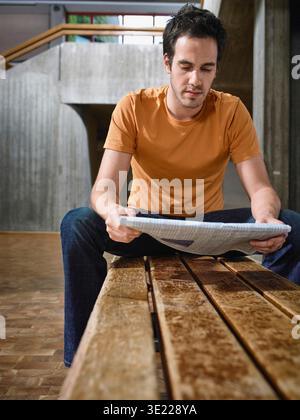 Un giovane con la camicia arancione si siede su una panchina e legge un giornale al chiuso. Germania Foto Stock