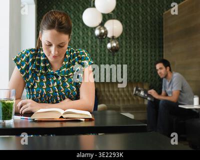 Donna che legge un libro a un tavolo in un bar moderno con un muro verde a motivi geometrici, uomo che guarda verso di lei. Germania Foto Stock
