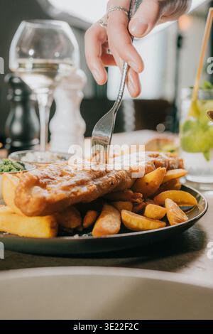 Solleva la forchetta sopra il pesce fritto dorato e le patatine fritte. Il piatto poggia su una superficie calda e rustica del tavolo. Un bicchiere di vino bianco si illumina delicatamente sullo sfondo. Cena Foto Stock