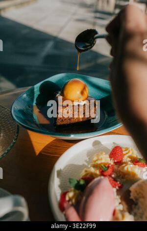 La luce calda del sole bagna un piatto da dessert. Versare a mano lo sciroppo su una torta dorata. Una piccola pesca si trova sulla torta. Le fragole e la panna si trovano accanto al Foto Stock