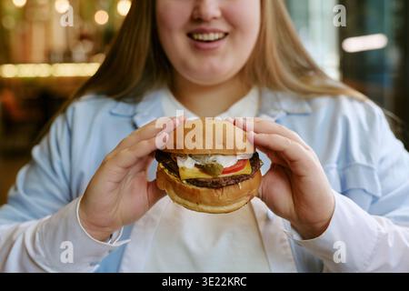 Giovane donna adulta sorridente mentre tiene l'hamburger in casa Foto Stock