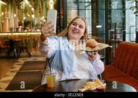 Giovane donna che prende Selfie mentre tiene un Burger al ristorante Foto Stock