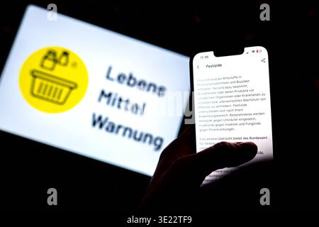 Brema, Germania. 17 marzo 2026. Una persona è in possesso di uno smartphone sul quale è aperta l'app "Food Warning" ed è possibile visualizzare sul display il testo informativo "pesticidi". Il logo dell'applicazione è visibile sullo sfondo. L'app fornisce informazioni sulle avvertenze correnti e sui richiami di prodotti per alimenti e altri prodotti di consumo in Germania. Credito: Sina Schuldt/dpa/Alamy Live News Foto Stock
