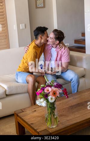 Uomo asiatico e partner maschio che condividono il bacio su un divano in pelle che tiene la scatola blu a casa Foto Stock