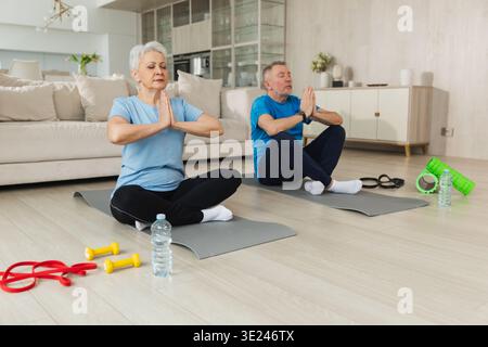 Meditazione yoga mindfulness. Coppia matura adulta anziana che pratica yoga a casa. Marito e moglie di mezza età seduti in posa di loto su un tappetino yoga per meditare e rilassarsi. Famiglia che fa pratica respiratoria Foto Stock
