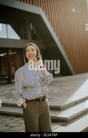 Una donna coreana si trova fuori da un edificio moderno. Sta sorridendo e parlando al telefono. Il sole splende su di lei mentre le piace la conversazione. Foto Stock