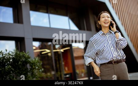 Una donna coreana si trova fuori da un edificio moderno. Sta sorridendo mentre parla al telefono. Il cielo è limpido, e ci sono grandi finestre dietro di lei. Foto Stock