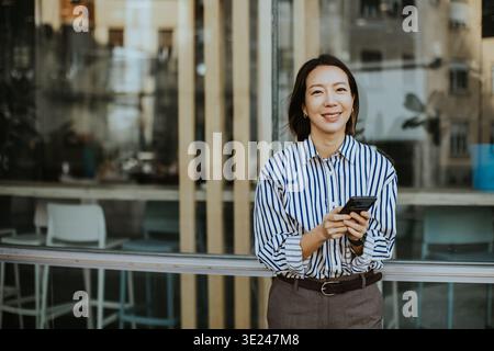 Una donna coreana sorride mentre tiene il suo smartphone. Si appoggia contro una ringhiera all'esterno di un edificio moderno. La scena la mostra in un abbigliamento casual Foto Stock
