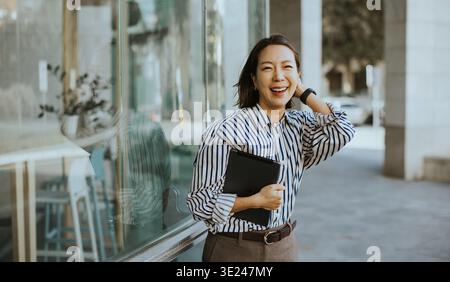 Una donna coreana ama trascorrere il proprio tempo fuori da un bar durante il giorno. Sta accanto a una finestra con un taccuino in mano, sorridente e apparente allegra. Lei Foto Stock