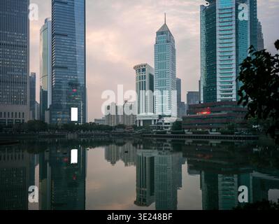Vista simmetrica di un moderno skyline metropolitano caratterizzato da alti grattacieli di vetro che si riflettono chiaramente in un tranquillo lago urbano durante il crepuscolo serale Foto Stock