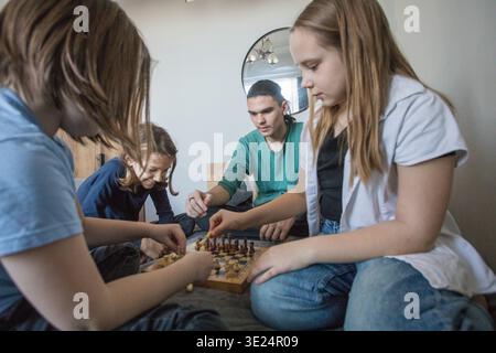 Quattro bambini si riuniscono in un soggiorno per giocare a scacchi su una tavola di legno. Si concentrano sul loro gioco mentre sono seduti su una superficie morbida. Il tempo passa come se fossero Foto Stock