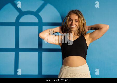 Giovane donna con dreadlock e tatuaggi che si posa felicemente contro un muro blu con l'ombra della finestra Foto Stock