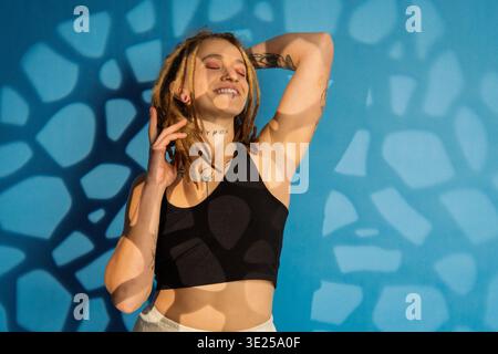 Giovane donna con dreadlock e tatuaggi in posa contro una parete blu con luce solare dappata Foto Stock
