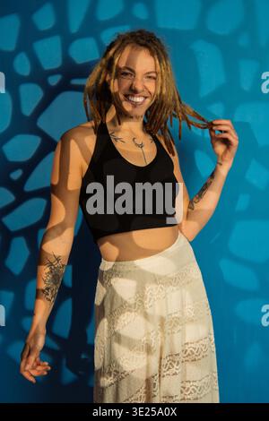 Giovane donna con dreadlock e tatuaggi sorridenti, in piedi in ombre fantasia su sfondo blu Foto Stock
