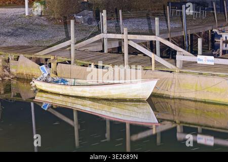 Una piccola barca bianca è ormeggiata lungo un molo in legno con acque calme che riflettono la barca e il molo. La scena è illuminata con un'illuminazione soffusa Foto Stock