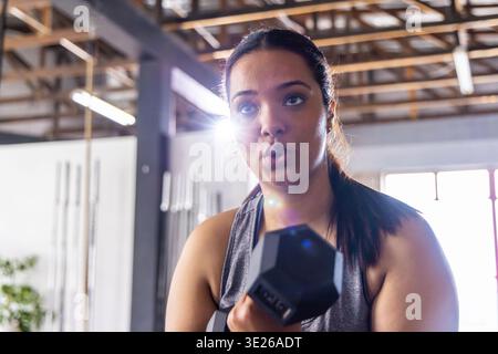 Donna indiana che solleva un manubrio esagonale vicino al petto in palestra con baracche, con la parte superiore grigio scuro Foto Stock