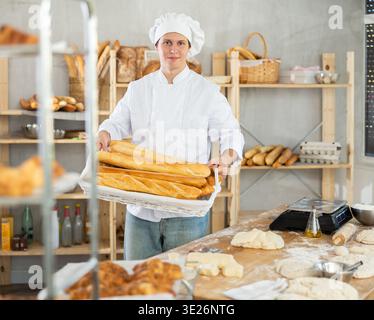 Giovane uomo positivo panettiere in uniforme bianca cuocere baguette fresche in panetteria Foto Stock