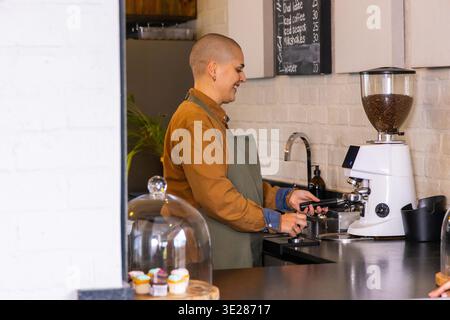 Barista maschile che indossa un grembiule verde per preparare l'espresso con portafiltro al bancone del caffè con cupcake Foto Stock