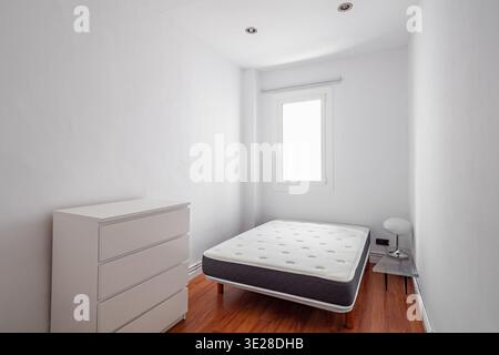 Camera minimalista con letto singolo e comò bianco. La sala ha pareti bianche e pavimento in legno, con una finestra che consente luce naturale. Foto Stock