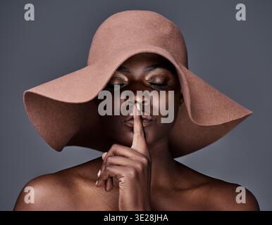 Cappello, donna nera e dita sulle labbra in studio, segreto di moda e bellezza per un gesto di silenzio. Privacy, notizie riservate e persona affrettata Foto Stock