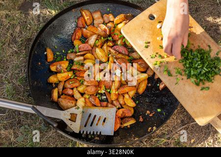 Patate da padella all'aperto con erbe fresche. Le patate arrostite dorate si cuocono in una grande teglia all'aperto, mentre le erbe fresche tritate vengono aggiunte su un legno Foto Stock