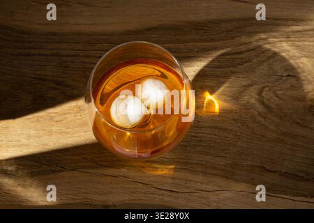 Un colpo ad alto contrasto di un bicchiere di whisky con grandi cubetti di ghiaccio su un rustico tavolo in legno. Un raggio di luce solare illumina il liquido color ambra Foto Stock