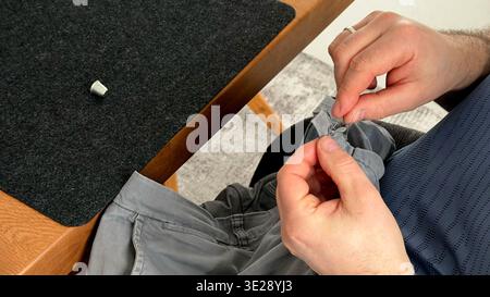 Uomo che cucisce bottone su tessuto grigio della camicia mentre è seduto a un tavolo di legno con superficie scura in feltro Foto Stock