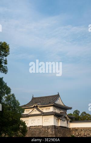 Storico castello giapponese arroccato su un muro di pietra sotto un morbido cielo blu, incorniciato da alberi verdi e da un aeroplano lontano. Architettura calma e senza tempo Foto Stock