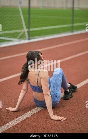 Seduta su una pista, la donna con l'attrezzatura sportiva blu prende una pausa, sottolineando l'importanza dei periodi di recupero nel mantenere la salute generale e. Foto Stock