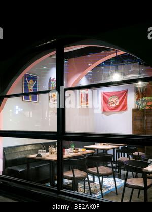 Vista notturna esterna che si affaccia su una sala da pranzo calda e luminosa del ristorante attraverso una grande finestra ad arco con cornice nera. In evidenza l'interio moderno Foto Stock