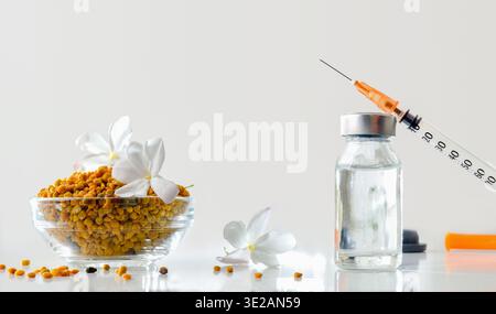 Concetto di vaccinazione antipolline con flaconcino di prodotto vaccinale e siringa riflessa su un tavolo bianco e una ciotola di vetro piena di allergeni. Vista frontale. Foto Stock