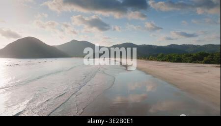 Selong Belanak Beach, Lombok: Incredibile e tranquilla baia tropicale con onde dolci e sabbia bagnata che rispecchia cielo blu e nuvole, incorniciata da lussureggianti colline verdi e costa lontana. Volo aereo con drone Foto Stock