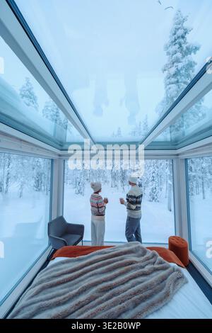 Annidato nel paesaggio innevato di Akaslompolo, due visitatori si godono bevande calde all'interno di una moderna cabina di vetro. La vista spettacolare degli alberi invernali all'esterno crea un'atmosfera serena perfetta per il relax. Foto Stock