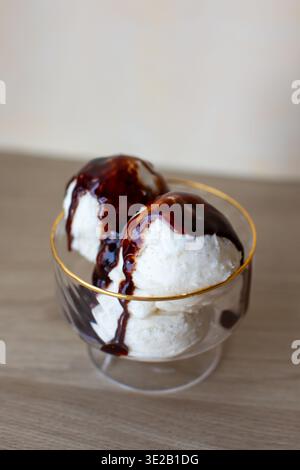 cucchiai di gelato ai fagioli di vaniglia serviti in una ciotola da dessert in vetro trasparente con bordo dorato. Il dessert è condito con ricco sciroppo di cioccolato fondente o fudge Foto Stock