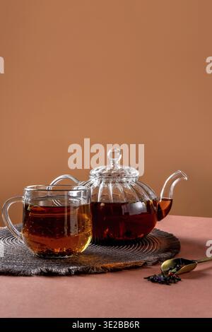 Teiera di vetro, tazza di tè e foglie di tè su sfondo marrone. Il tè alle erbe caldo è nella teiera di vetro sul tavolo. Copia spazio Foto Stock