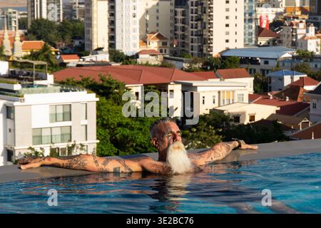 Uomo tatuato con lunga barba bianca che si rilassa in una piscina a sfioro sul tetto affacciata sul paesaggio urbano di Phnom Penh, Cambogia. Foto Stock