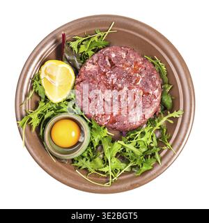Vista dall'alto della porzione di tartare di bistecca (carne di manzo macinata cruda e tuorlo crudo nel recipiente su verdure fresche) su piatto marrone isolato su sfondo bianco Foto Stock