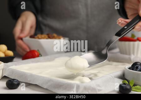 Preparare la corteccia dello yogurt. Donna che spalma lo yogurt con un cucchiaio sul piatto a tavola bianca, primo piano Foto Stock