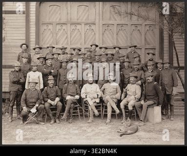 Fotografia seppia di ufficiali Rough Rider che mostrano Theodore Roosevelt seduto tra colleghi ufficiali con un leone di montagna ai suoi piedi, circondato da truppe in piedi in uniforme militare poste di fronte a un edificio in legno. Foto Stock