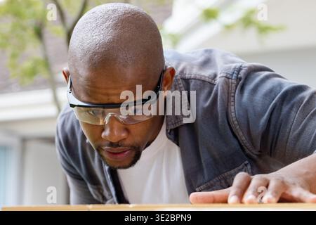 Uomo afroamericano che si appoggia, ispeziona la tavola di legno sul portico della casa indossando occhiali di sicurezza. Lavoratore di legno, banco da lavoro, tavola, occhiali, occhiali protettivi Foto Stock